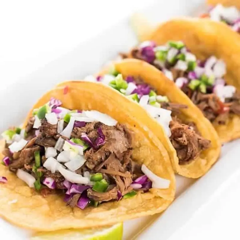 A slow-cooked beef taco inspired by the savory flavors of ropa vieja. Tender shredded beef simmered in a tomato-based sauce with garlic, onions, and bell peppers piled into a warm, soft tortilla and topped with fresh ingredients like crisp lettuce, diced onions, cilantro, and a squeeze of lime. at The Food Corner | Toast
